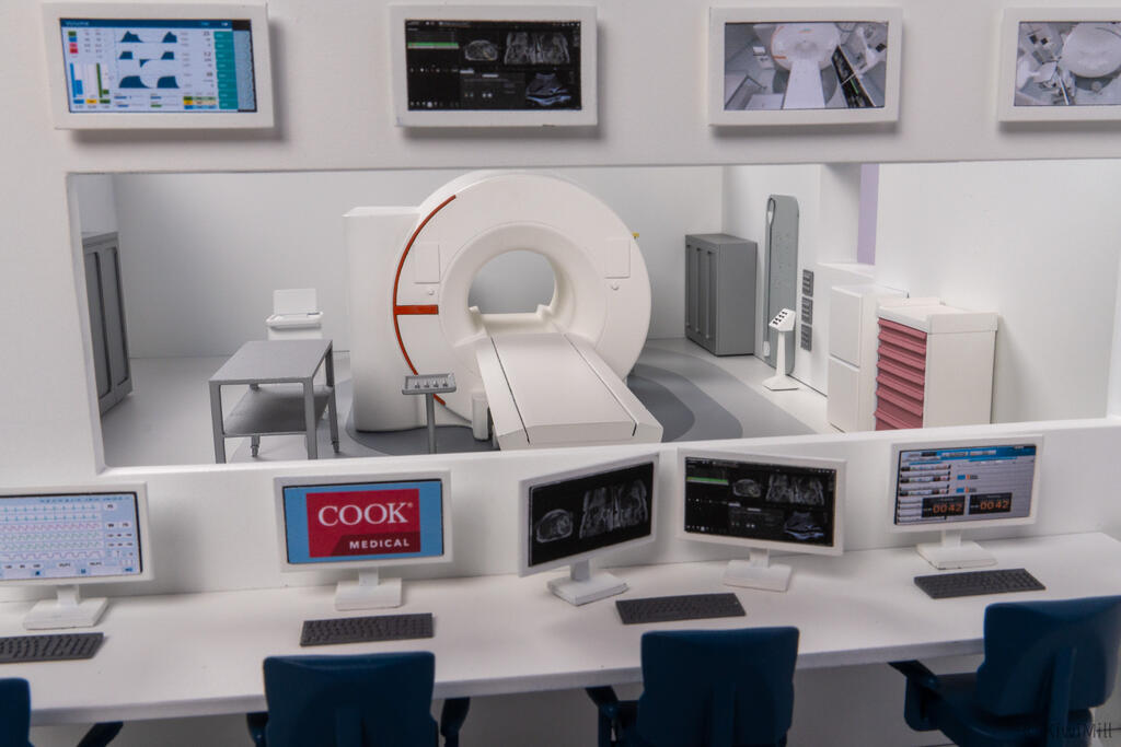 MRI suite scale model viewed through the control room window, with workstations, wall displays and scanner beyond